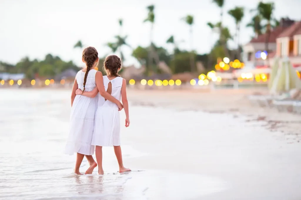 Two children in white dresses holding each other while walking on the beach