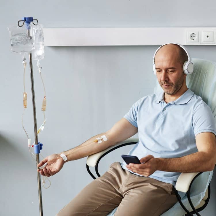Bald adult man using smartphone during IV drip treatment in clinic