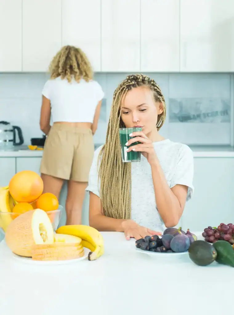 Young woman drinking green smoothie in modern kitchen with fruits.