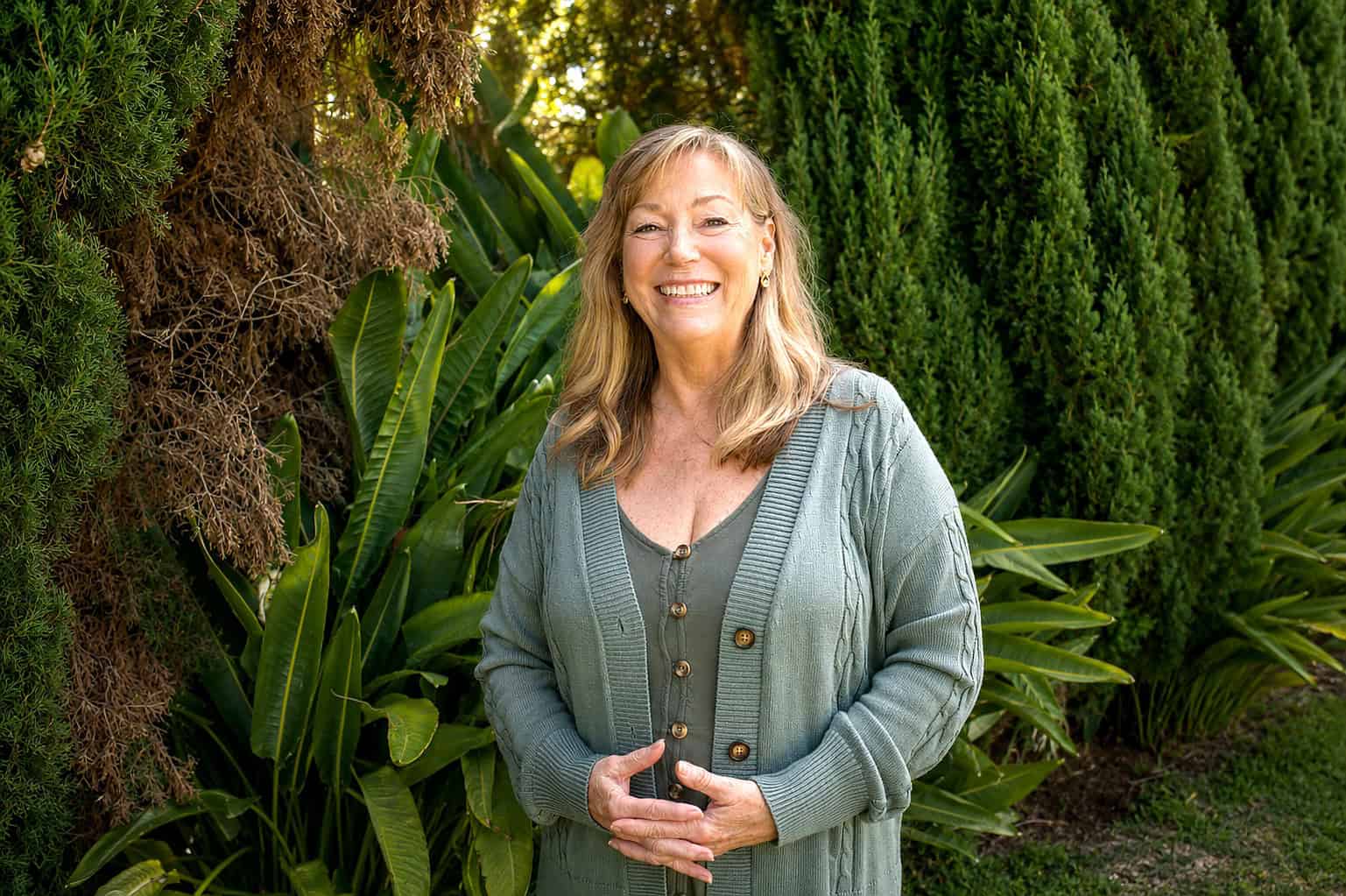 Smiling woman standing outdoors among lush green plants and trees.
